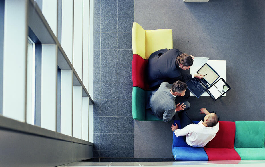 Three business people in discussion sitting on colourful seats, overhead view.